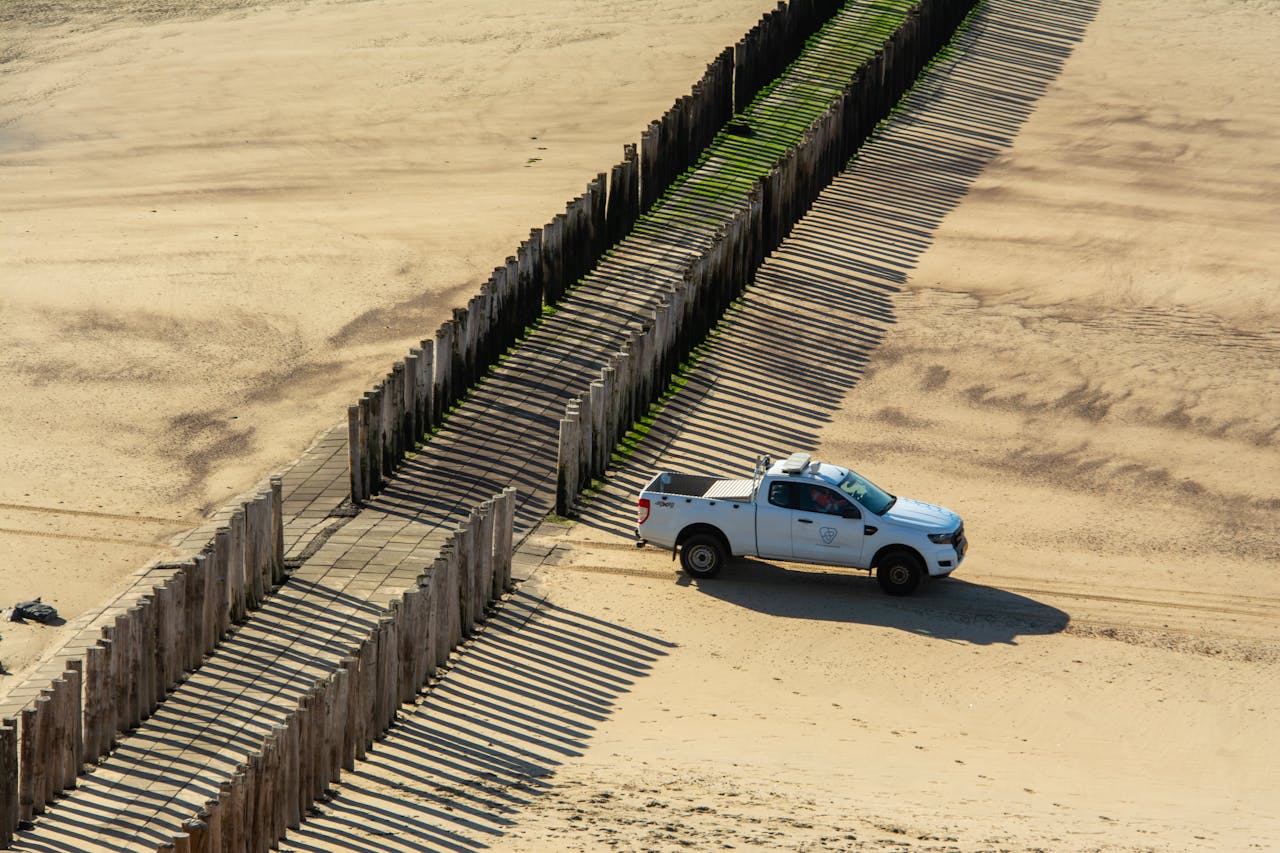 Aerial shot of a white pickup truck driving along a sandy beach beside wooden pilings casting long shadows.