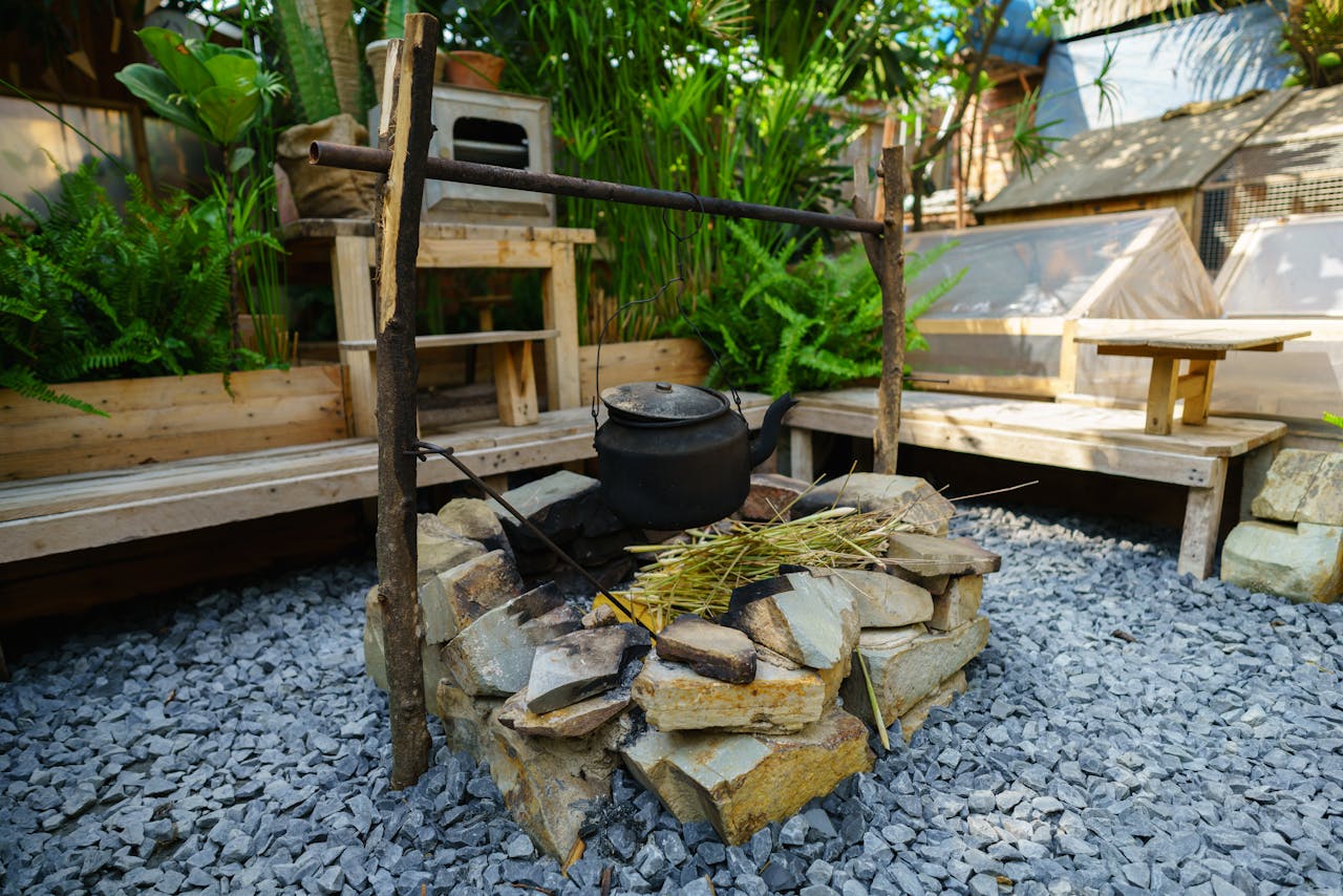 Cozy outdoor cooking area with a stone firepit and vintage pot, set in a lush garden environment.