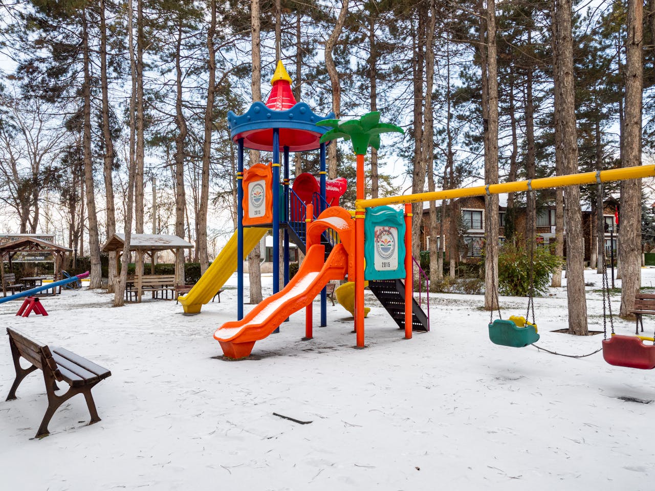 Bright playground equipment stands in a snowy park surrounded by bare trees, capturing a serene winter day.