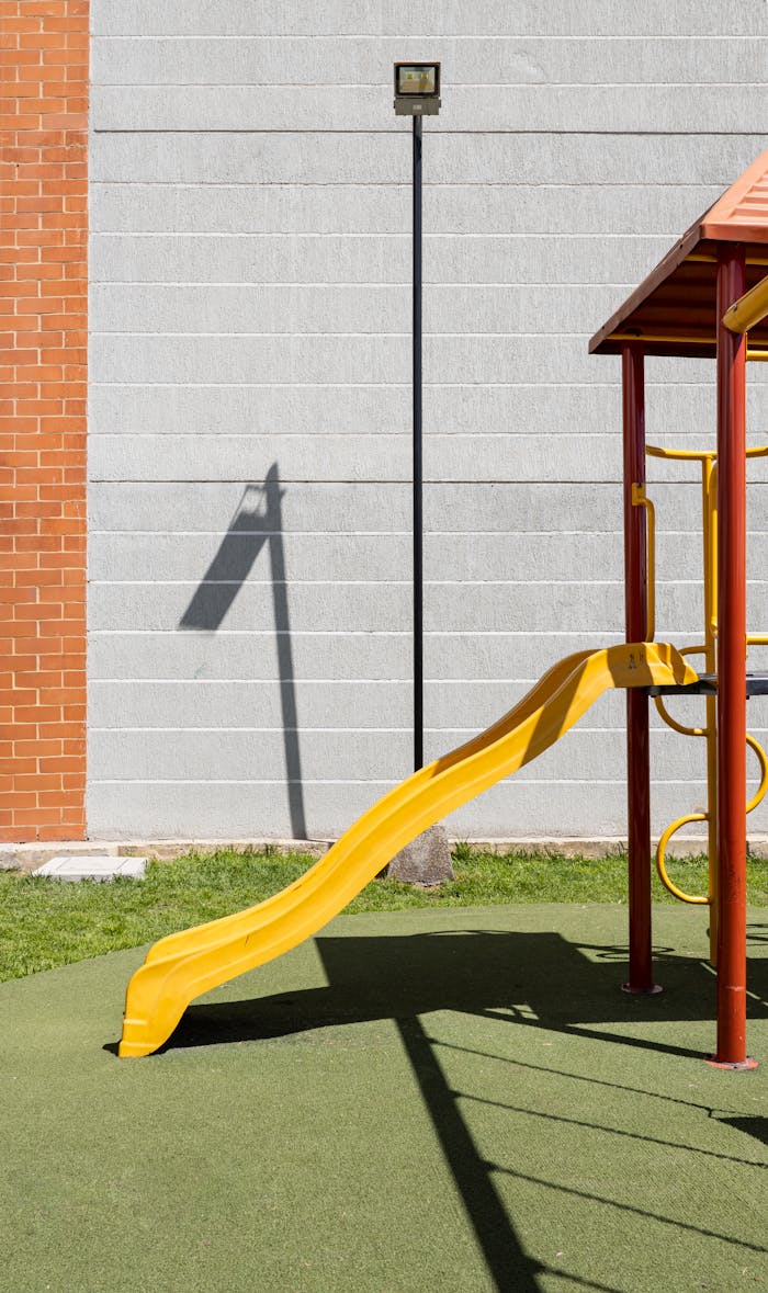 A sunny day in a park with an empty yellow slide casting a shadow on green turf.